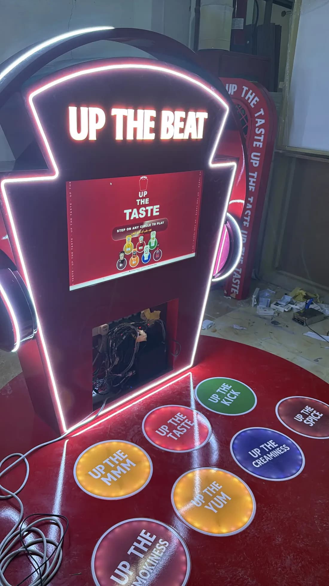 The built Up The Beat kiosk in the workshop: a neon-edged Heinz keystone wearing headphones, its screen reading “Up the Taste, step on any circle to play” in English and Arabic, with seven lit floor circles in front of it.
